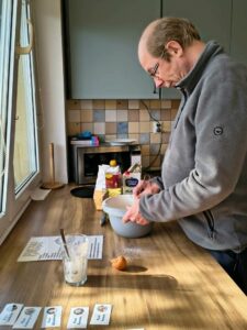 Mężczyzna w okularach stojący przy blacie kuchennym. Miesza w misce składniki na ciasto. Na blacie stoi mąka, skorupki po jajkach, karton mleka.