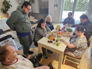 Trzy kobiety i dwóch mężczyzn siedzi przy stole i obierają i kroją owoce. Usmiechają się. Jeden mężczyzna stoi obok i przygląda się. Kolejny mężczyzna siedzi obok na wózku inwalidzkim.